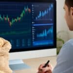 A commodities trader analyzing sesame market trends on a computer screen displaying price charts and trading graphs, with sacks of sesame seeds placed on the desk in the foreground.
